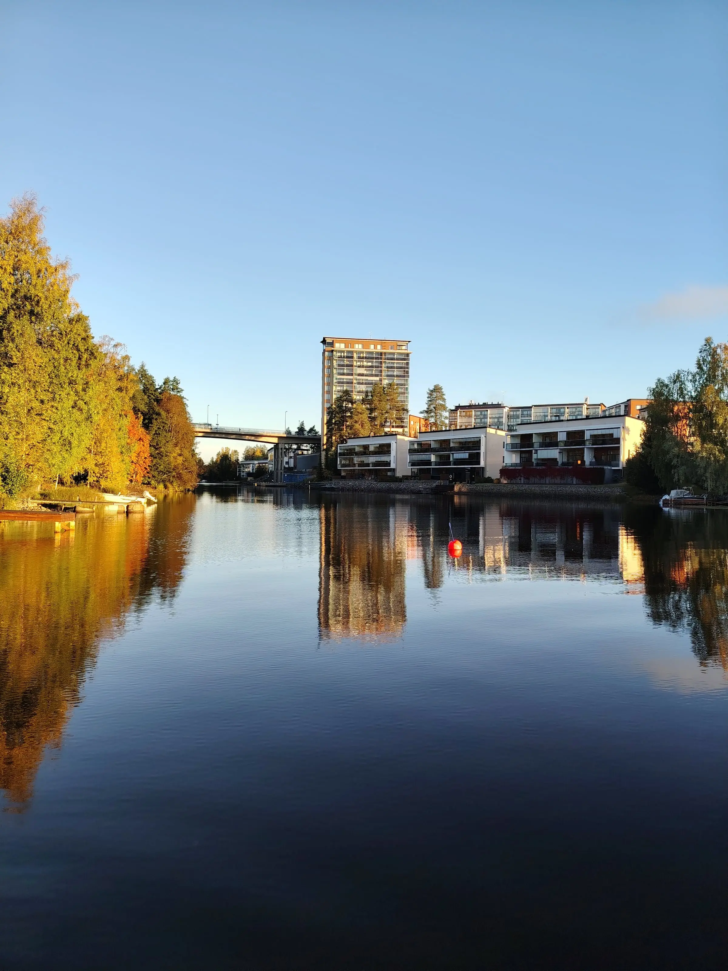 The still lake reflecting autumn trees and blocks of flats on the far shore.